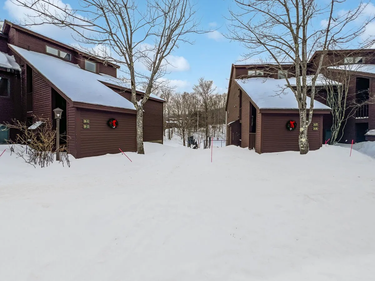 A snow-covered exterior showing two brown houses with holiday wreaths, bare trees, and ample open space. The scene highlights direct trail access and abundant parking, with a clear path for easy coming and going.