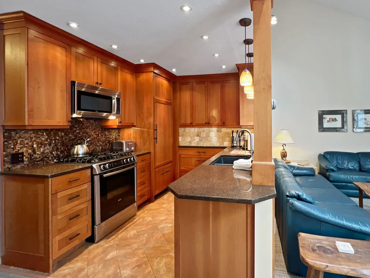 This image showcases a stylish, fully renovated kitchen with stainless steel appliances, rich wood cabinetry, and a gleaming granite countertop—seamlessly blending into a cozy living area with a plush blue leather sofa. The space is warm, inviting, and perfect for relaxing after a day on the slopes or gathering for family meals.