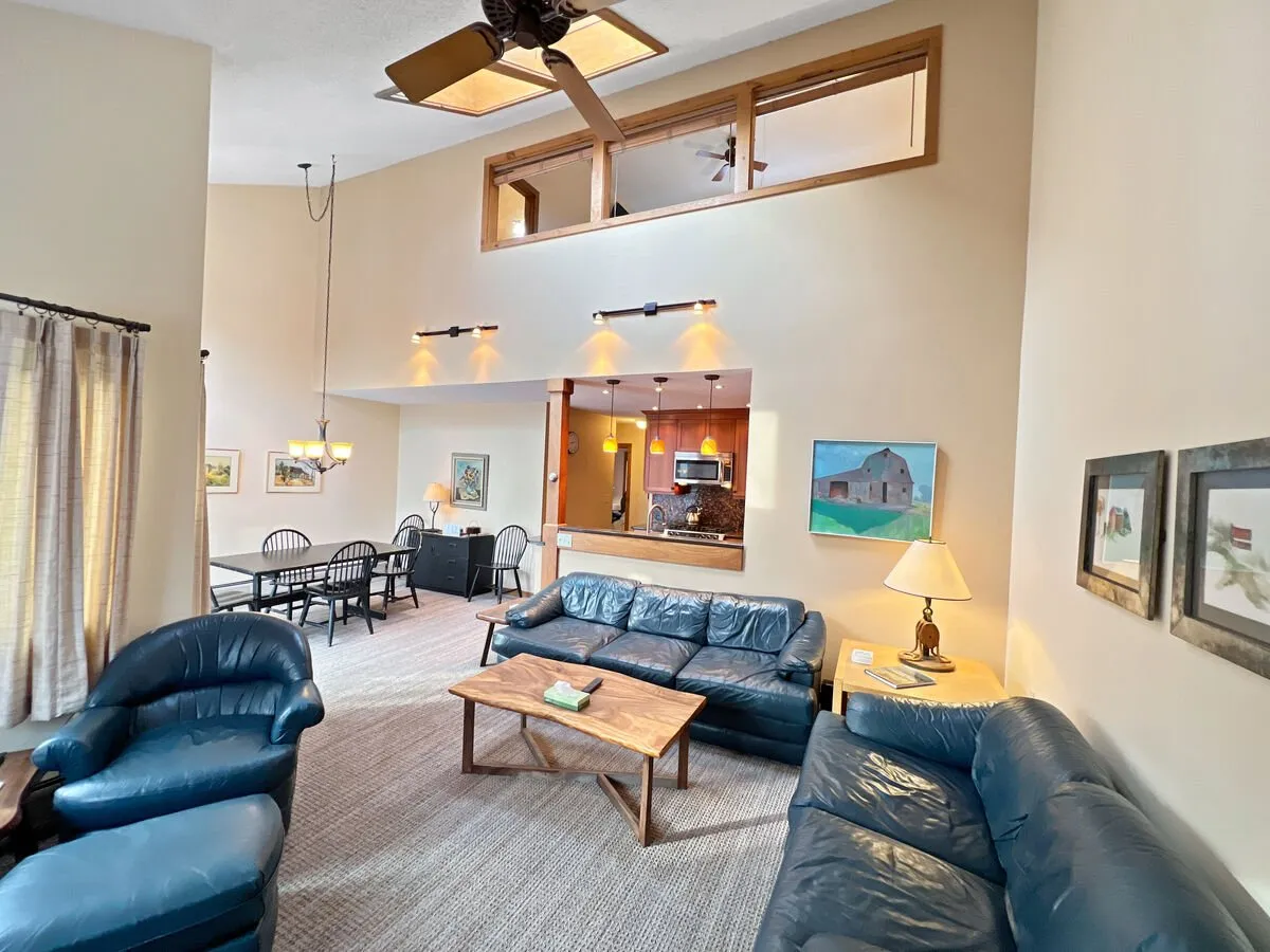 A wide-angle view of the open-plan living, dining, and kitchen spaces. The leather sofas and wooden coffee table invite gatherings, while the adjoining dining area and kitchen opening emphasize the seamless flow between spaces. Sunlight streams through the windows, enhancing the inviting and social atmosphere.