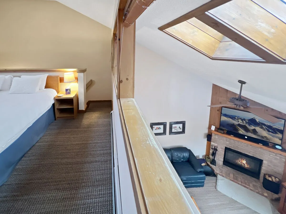 A welcoming living area with white-linen bed, rustic fireplace with a wood mantle, leather couch, and abundant natural light streaming through skylights. The scene is warm and inviting, ideal for relaxing, socializing, or cozying up by the fire.