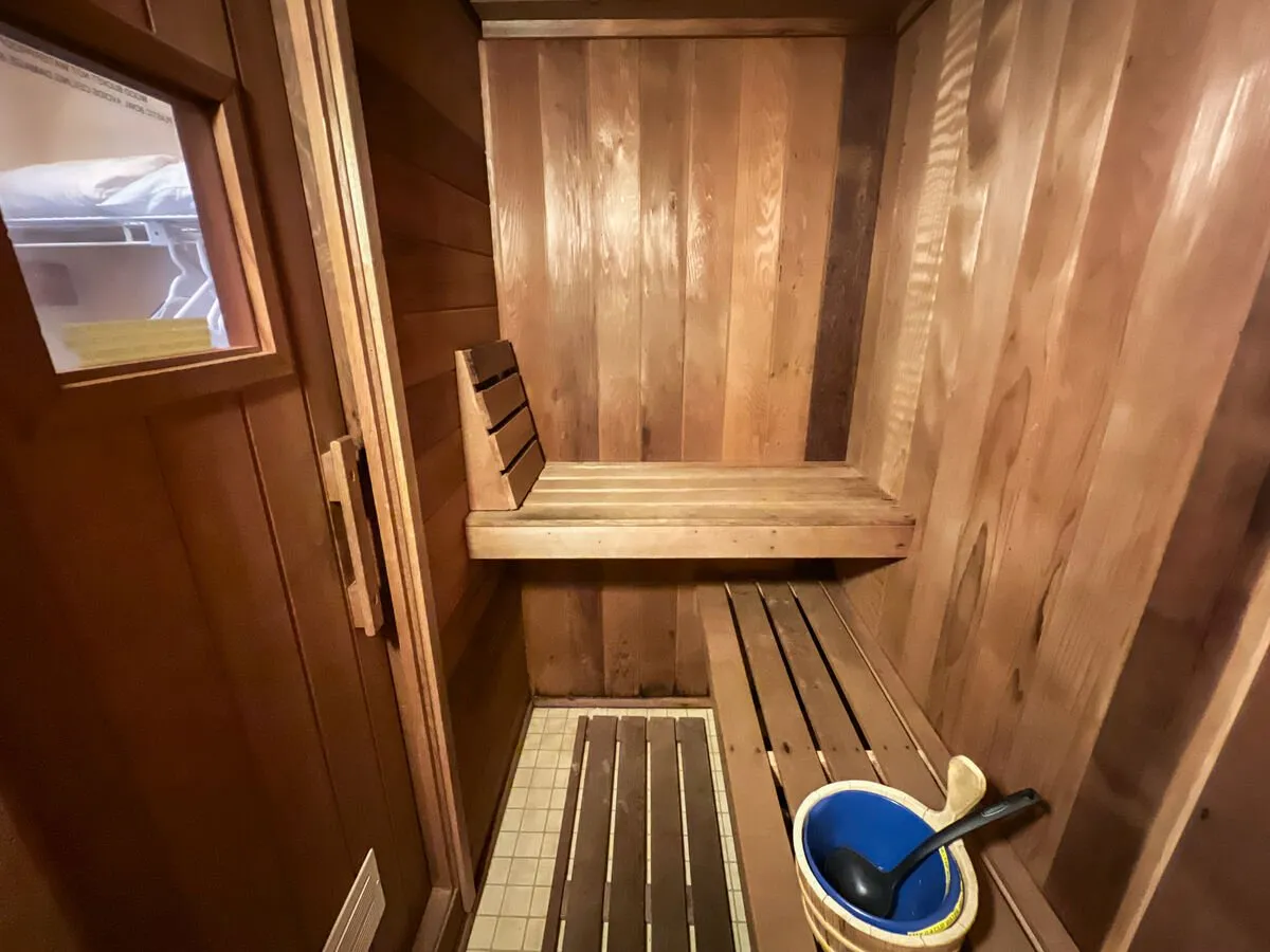 A cozy, wood-paneled sauna featuring wooden benches, a small window letting in soft light, and a classic sauna bucket with ladle. The tranquil ambiance and intimate scale evoke relaxation and indulgence, aligning perfectly with the promise of private après-ski comforts.