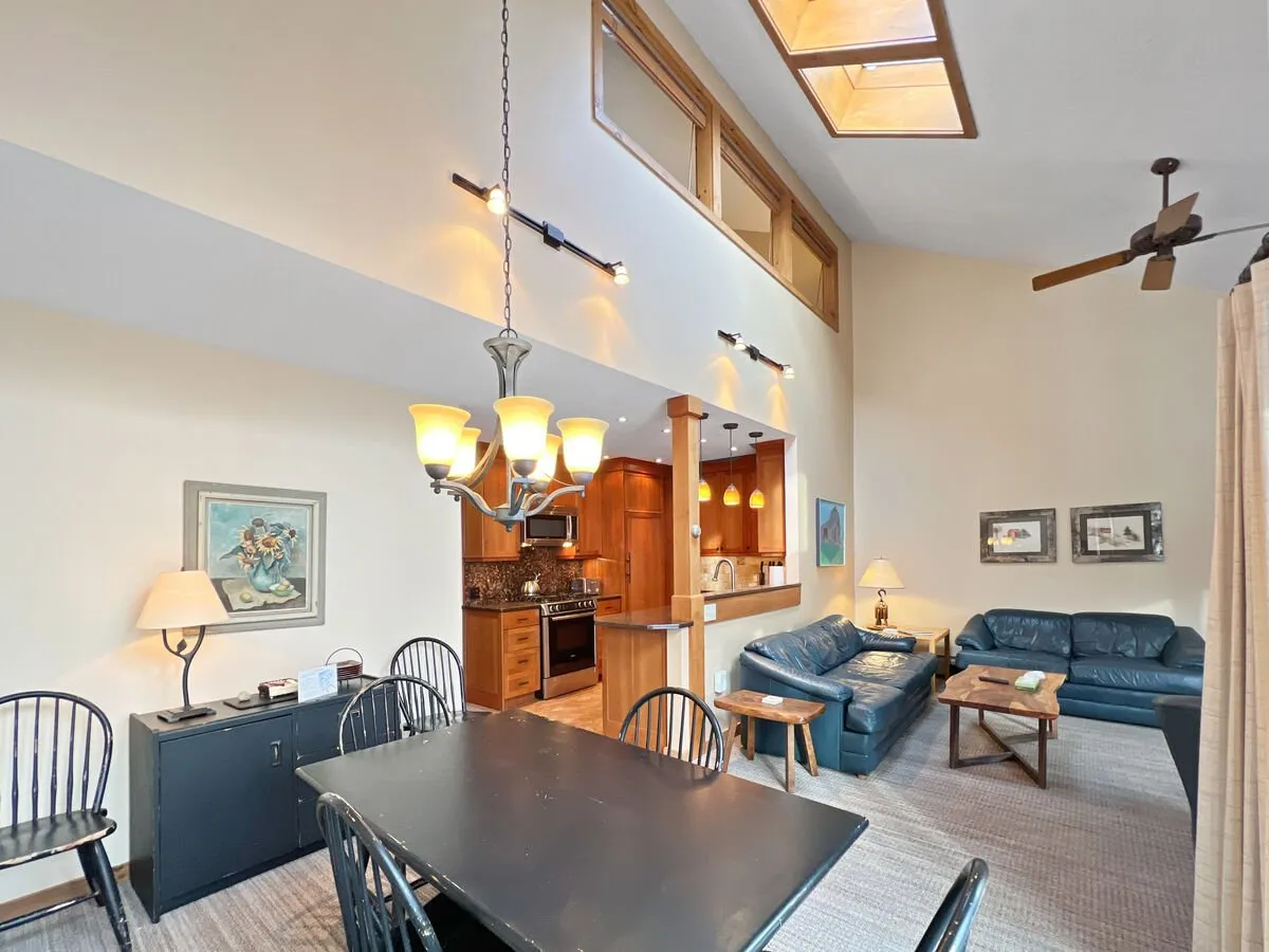 This image presents an open-concept interior where the dining table sits adjacent to the kitchen area. The space is warmly lit by a central chandelier, with the living room seating visible in the background. The kitchen's cabinetry and appliances are partially visible, hinting at the custom craftsmanship and high-end finishes. The inviting setup suggests a hub for family meals, social gatherings, or relaxed evenings.