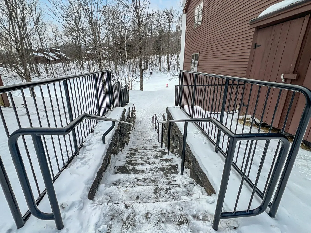 An exterior view capturing the snow-covered ground, stairway with handrail, surrounding trees, and the building's entrance. The atmosphere is calm and wintry, suggesting easy access to outdoor adventures right from the doorstep.