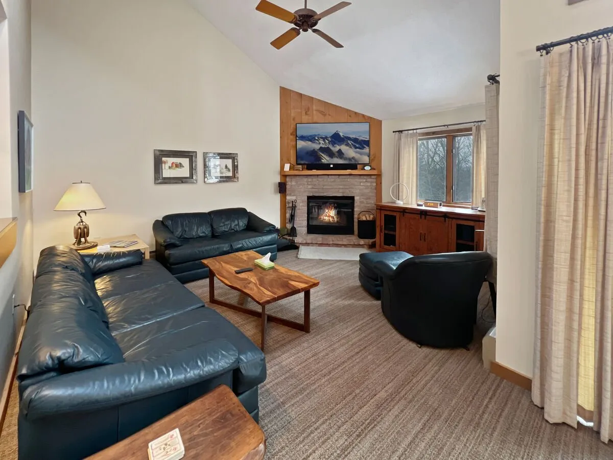 This image captures a spacious living room featuring rich leather sofas arranged around a wooden coffee table. A prominent wood-burning fireplace and a large OLED Smart TV are focal points, with a scenic mountain view visible through the window. The overall atmosphere is warm, ideal for relaxation or movie nights after skiing.