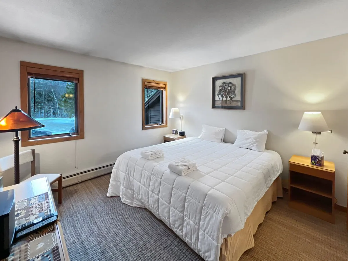 A bright and calm bedroom with a king-sized bed adorned with white bedding, natural light streaming through the window, tasteful wall art, and a bedside lamp. The space feels restful and inviting.