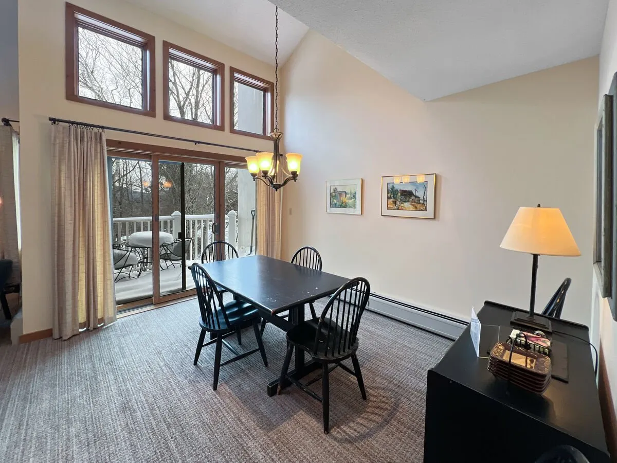 A cozy dining area with a wooden table set beneath a stylish chandelier, flanked by artwork on the walls. Sliding glass doors open to mountain views, suggesting a space ideal for sharing meals and stories in a warm, inviting setting.