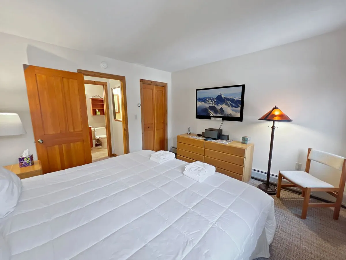 A cozy bedroom scene with a queen bed accented by neatly arranged towels, a wooden dresser, a television displaying a scenic mountain landscape, and a soft table lamp. The setup suggests a relaxing space for watching TV or unwinding after group activities.